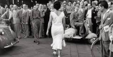 Una signora vestita di bianco, risperesa di spalle, mentre cammina sotto gli sguardi di un gruppo di uomini