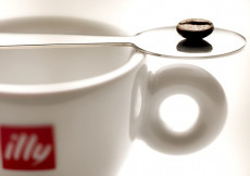Un chicco di caffè posato su un cucchiaino sopra una tazzina con il logo di Illy