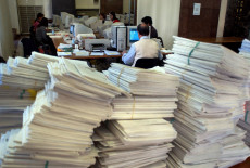 Faldoni di documenti accatastati su un tavolo di un ufficio della Pubblica Amministrazione.